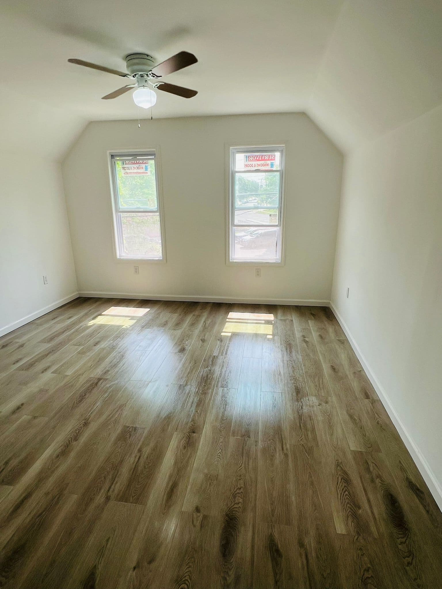 Sunlit empty room featuring polished wood floors, two windows, and a white ceiling fan.